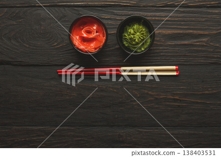 Pieces of traditional japanese tablewear. Red chopsticks with hierogliphs, small black bowls with ginger and wasabi on dark wooden table. Top view, flat lay, copy space, background Pieces of traditional japanese tablewear. Red chopsticks with hierogliphs, small black bowls with ginger and wasabi on dark wooden table. Top view, flat lay, copy space, background 138403531