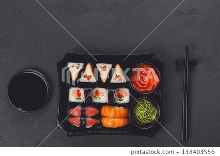 Japanese food restaurant delivery, take away box, sushi maki and roll set. Chopsticks, ginger, soy sauce, wasabi. Meals at black background, top view, copy space Japanese food restaurant delivery, take away box, sushi maki and roll set. Chopsticks, ginger, soy sauce, wasabi. Meals at black background, top view, copy space 138403556