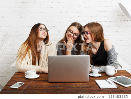 Three friends sit around a table with a laptop, sharing laughter and conversation. Two cups are placed on the table. The setting is bright and inviting with a white brick wall. 138403585