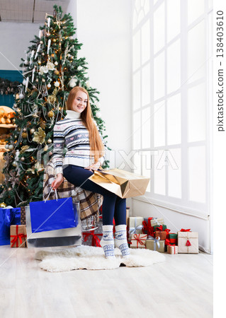 Happy girl with packages after christmas shopping. Smiling young woman in casual at home interior with decorated xmas tree and gift boxes posing on camera. Winter holidays sales. Copy spase Happy girl with packages after christmas shopping. Smiling young woman in casual at home interior with decorated xmas tree and gift boxes posing on camera. Winter holidays sales. Copy spase 138403610