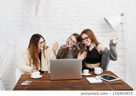 Three friends are sitting at a wooden table, smiling and waving at the laptop. They have drinks and items on the table. The room has white walls and a lamp, creating a cheerful scene. Three friends are sitting at a wooden table, smiling and waving at the laptop. They have drinks and items on the table. The room has white walls and a lamp, creating a cheerful scene. 138403655