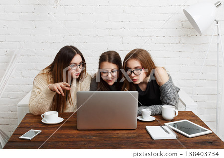 Three friends sit at a wooden table with coffee cups, looking intently at a laptop screen. They are engaged in conversation and sharing ideas in a cozy workspace. Three friends sit at a wooden table with coffee cups, looking intently at a laptop screen. They are engaged in conversation and sharing ideas in a cozy workspace. 138403734
