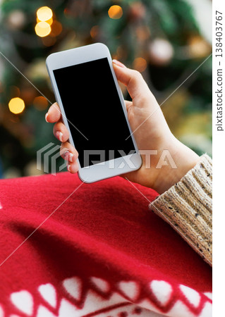 Christmas online shopping. Female buyer makes order at screen of smartphone with copy space. Woman buys presents for xmas eve, sits at home near pine tree with shining lights. Winter holidays sales 138403767