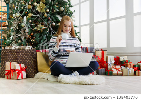 Young woman with coffee shopping online on laptop in christmas interior. Girl sitting under decorated fir tree among lots of wrapped presents. Preparing to xmas, bying on winter sales, copy space Young woman with coffee shopping online on laptop in christmas interior. Girl sitting under decorated fir tree among lots of wrapped presents. Preparing to xmas, bying on winter sales, copy space 138403776