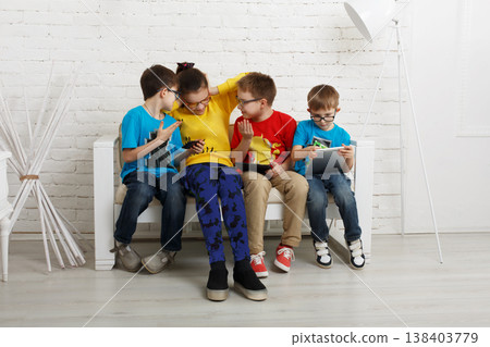 Four children are sitting on a couch in a bright room. They are engaged in games and chatting with each other. The atmosphere is lively as they share their excitement. 138403779