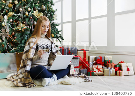 Young woman with credit card shopping online on laptop in cozy christmas interior. Girl sitting under decorated pine tree among wrapped presents. Preparing to xmas, bying on winter sales, copy space 138403780