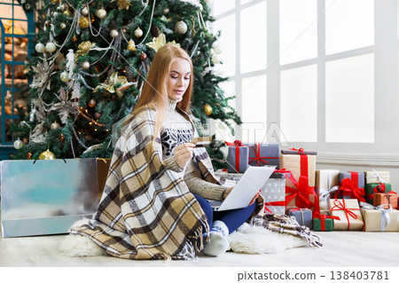 Young woman with cradit card shopping online on laptop in christmas interior. Girl sitting under decorated fir tree among lots of wrapped presents. Preparing to xmas, bying on winter sales. Copy space 138403781