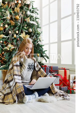 Young woman talking on mobile and shopping online on laptop in cozy christmas interior. Girl sit under decorated pine tree among wrapped presents. Preparing to xmas, bying on winter sales, copy space Young woman talking on mobile and shopping online on laptop in cozy christmas interior. Girl sit under decorated pine tree among wrapped presents. Preparing to xmas, bying on winter sales, copy space 138403792