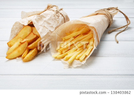 Potato wedges and french fries wrapped in brown wrapping paper. Fast food take away at white shabby chic wood. Fried potatoes with tomato sauce. Chips, potato slices. Top view, flat lay, closeup. Potato wedges and french fries wrapped in brown wrapping paper. Fast food take away at white shabby chic wood. Fried potatoes with tomato sauce. Chips, potato slices. Top view, flat lay, closeup. 138403862