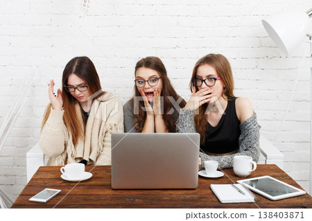 Three women sit at a wooden table with drinks, focused on a laptop. They show surprise and interest while watching something on the screen in a well-lit space with white walls. Three women sit at a wooden table with drinks, focused on a laptop. They show surprise and interest while watching something on the screen in a well-lit space with white walls. 138403871