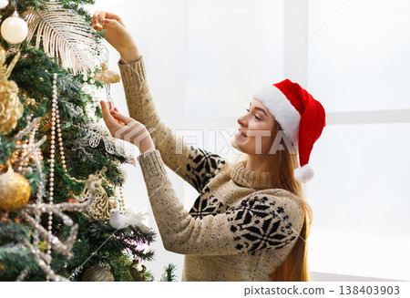 Young beautiful woman in santa hat decorating christmas tree. Stylish modern design, golden baubles, white beads and shiny garland. Winter holidays background with copy space 138403903