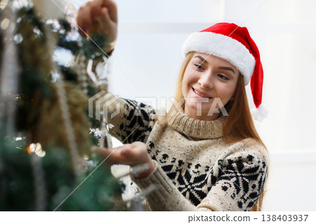 Young beautiful woman in santa hat decorating christmas tree. Stylish modern design, golden baubles, white beads and shiny garland. Winter holidays background with copy space Young beautiful woman in santa hat decorating christmas tree. Stylish modern design, golden baubles, white beads and shiny garland. Winter holidays background with copy space 138403937