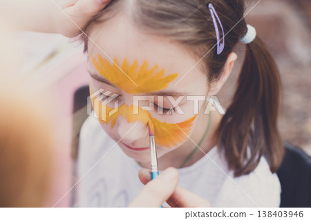 Child animator, artist hand draws face painting to little girl. Child with funny face painting. Painter makes tiger eyes at girl face. Children holiday, event, birthday party, entertainment. Child animator, artist hand draws face painting to little girl. Child with funny face painting. Painter makes tiger eyes at girl face. Children holiday, event, birthday party, entertainment. 138403946