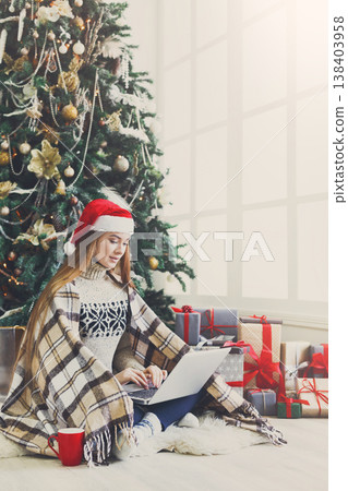 Girl with laptop sitting on the floor at decorated christmas tree background. Young woman writing greeting emails online in holiday home atmosphere among present boxes. Copy space on window 138403958