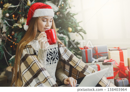 Girl with laptop sitting on the floor at decorated christmas tree background. Young woman drinking coffee and writing greeting emails online in holiday home atmosphere among present boxes. Copy space Girl with laptop sitting on the floor at decorated christmas tree background. Young woman drinking coffee and writing greeting emails online in holiday home atmosphere among present boxes. Copy space 138403964