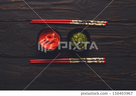 Pieces of traditional japanese tablewear. Red chopsticks with hierogliphs, small black bowls with ginger and wasabi on dark wooden table. Top view, flat lay, copy space, background 138403967