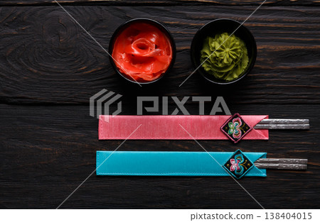 Pieces of traditional japanese tablewear. Chopsticks in blue and pink cases, small black bowls with ginger and wasabi on dark wooden table. Top view, flat lay, copy space, background 138404015