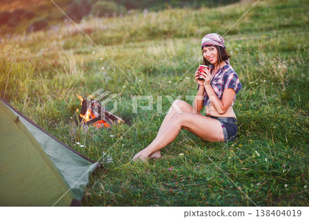 Girl near bonfire. Travel, tourism, camping - young slim sporty woman tourist brunette at the beautiful nature landscape sitting near the tent by the fire. Girl with drink in mug in her hands. 138404019