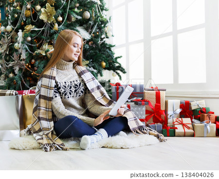 Christmas online shopping. Female buyer choosing presents on digital tablet sitting at decorated fir tree background among gift boxes and packages. Winter sales, copy space 138404026