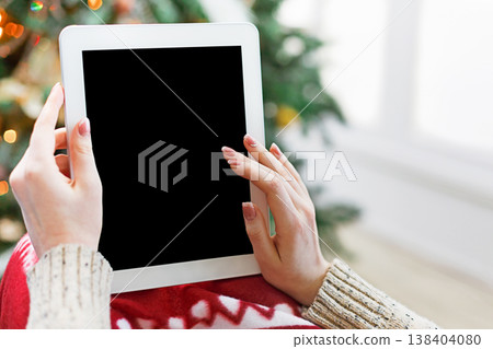 Christmas online shopping. Female buyer makes order at screen of tablet with copy space. Woman buys presents for xmas eve, sits at home near pine tree with shining lights. Winter holidays sales Christmas online shopping. Female buyer makes order at screen of tablet with copy space. Woman buys presents for xmas eve, sits at home near pine tree with shining lights. Winter holidays sales 138404080