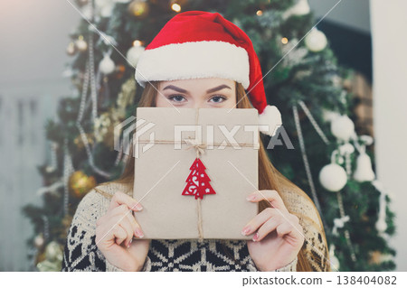 Young happy girl in red santa hat covering half face with christmas gift on xmas tree background. Funny and joyful winter holiday wallpaper Young happy girl in red santa hat covering half face with christmas gift on xmas tree background. Funny and joyful winter holiday wallpaper 138404082