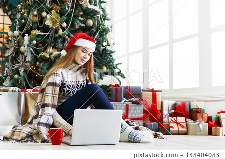 Girl with laptop sitting on the floor at decorated christmas tree background. Young woman drinking coffee, writing greeting emails online in holiday home atmosphere among present boxes. Copy space Girl with laptop sitting on the floor at decorated christmas tree background. Young woman drinking coffee, writing greeting emails online in holiday home atmosphere among present boxes. Copy space 138404083
