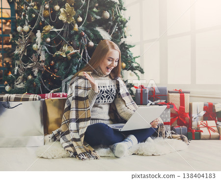 Surprised girl with laptop sitting at christmas tree background. Happy woman got greeting email from online shop with huge discount at winter sale. Holiday home with among present boxes, copy space 138404183