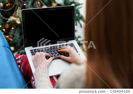 Christmas online shopping. Female buyer makes order at empty screen of laptop with credit card. Woman buys presents for xmas eve, sits at home near fir tree with shining lights. Winter holidays sales Christmas online shopping. Female buyer makes order at empty screen of laptop with credit card. Woman buys presents for xmas eve, sits at home near fir tree with shining lights. Winter holidays sales 138404189