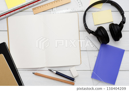 Stationery supplies in modern office workspace. Top view on open blank notebook, pen, pencil, rulers, small notepads and memo pads, headphones on white wooden desktop, flat lay, copy space, mockup 138404209