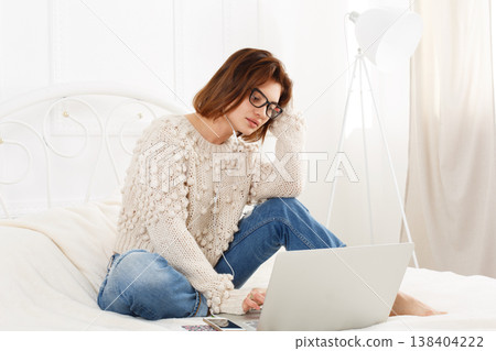 Young woman reads from laptop computer in bed. Engrossed Girl in eyeglasses and earphones with mobile phone. Attractive woman in casual clothes at white bed. Internet freelancer. High key, soft tone Young woman reads from laptop computer in bed. Engrossed Girl in eyeglasses and earphones with mobile phone. Attractive woman in casual clothes at white bed. Internet freelancer. High key, soft tone 138404222