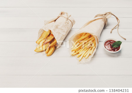 Potato wedges and french fries wrapped in brown wrapping paper. Fast food take away at white shabby chic wood. Fried potatoes with tomato sauce. Chips, potato slices. Top view, flat lay with copyspace Potato wedges and french fries wrapped in brown wrapping paper. Fast food take away at white shabby chic wood. Fried potatoes with tomato sauce. Chips, potato slices. Top view, flat lay with copyspace 138404231