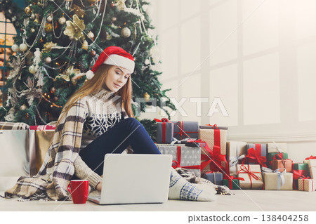 Girl with laptop sitting on the floor at decorated christmas tree background. Young woman drinking coffee and writing greeting emails online in holiday home atmosphere among present boxes. Copy space 138404258