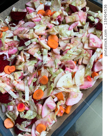 Vegetables including carrots, beets, and garlic are prepared on a baking tray ready to bake in the oven. The tray is lined and set on a stove surface 138404536