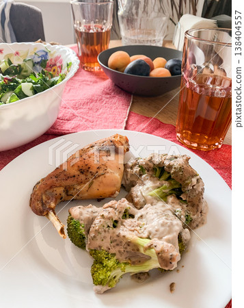 A plate features baked rabbit and broccoli with sauce. A fresh green salad sits nearby. Glasses of tea are filled. Apricots and plums are on the table for dessert A plate features baked rabbit and broccoli with sauce. A fresh green salad sits nearby. Glasses of tea are filled. Apricots and plums are on the table for dessert 138404557