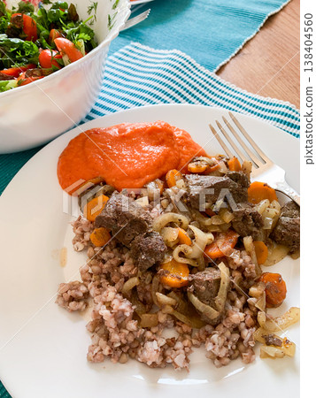 A plate holds beef liver with green buckwheat and squash caviar, alongside a fresh salad in a bowl. The table has a blue cloth adding color to the meal setting 138404560