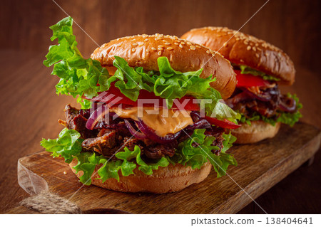 Two burgers with torn beef, tomatoes, fried onions and sauce, laid out on a wooden board, there are no people. Two burgers with torn beef, tomatoes, fried onions and sauce, laid out on a wooden board, there are no people. 138404641