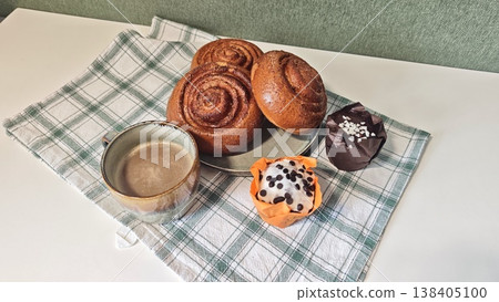 Freshly baked cinnamon rolls and a cupcake on a checkered tablecloth with a cup of coffee, creating a cozy breakfast setting in a modern kitchen Freshly baked cinnamon rolls and a cupcake on a checkered tablecloth with a cup of coffee, creating a cozy breakfast setting in a modern kitchen 138405100