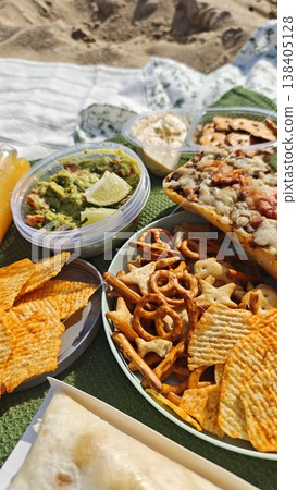 A variety of snacks such as guacamole, chips, pretzels, and a sandwich displayed on a picnic blanket at the beach with sand visible in the background A variety of snacks such as guacamole, chips, pretzels, and a sandwich displayed on a picnic blanket at the beach with sand visible in the background 138405128