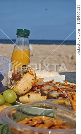 Delicious beach picnic featuring a loaded hot dog, crispy snacks, fresh grapes, and a bottle of orange juice against a scenic ocean backdrop Delicious beach picnic featuring a loaded hot dog, crispy snacks, fresh grapes, and a bottle of orange juice against a scenic ocean backdrop 138405135