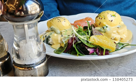 Plate of eggs benedict with hollandaise sauce, smoked salmon, and mixed greens served on a white dish with a pepper shaker in a restaurant setting Plate of eggs benedict with hollandaise sauce, smoked salmon, and mixed greens served on a white dish with a pepper shaker in a restaurant setting 138405273