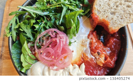 Dish includes poached egg in tomato sauce, garnished with fresh greens and pickled onions, accompanied by a slice of bread on a wooden surface Dish includes poached egg in tomato sauce, garnished with fresh greens and pickled onions, accompanied by a slice of bread on a wooden surface 138405274