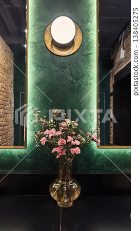 Floral arrangement of pink flowers in a clear vase placed on a black table against a green textured wall with a round wall light fixture Floral arrangement of pink flowers in a clear vase placed on a black table against a green textured wall with a round wall light fixture 138405275