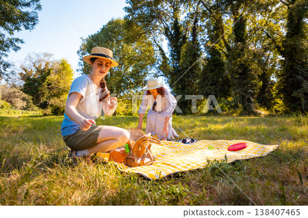Young daughter and her Caucasian mother sitting in a sunny park on a picnic blanket enjoying quality time, games, and family holiday outdoors. Low angle view Young daughter and her Caucasian mother sitting in a sunny park on a picnic blanket enjoying quality time, games, and family holiday outdoors. Low angle view 138407465