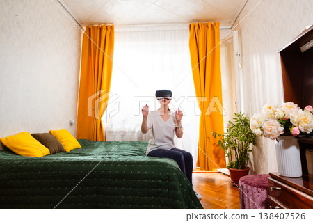 Woman wearing virtual reality headset while relaxing on bed in stylish bedroom, experiencing futuristic interactive media. Wide shot 138407526