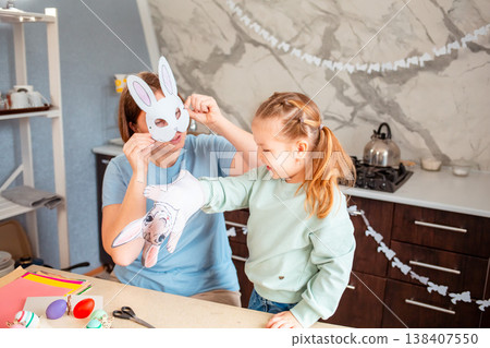 A happy Easter scene with a mother and daughter putting on paper bunny masks at the kitchen table while playfully preparing for the holiday together. A happy Easter scene with a mother and daughter putting on paper bunny masks at the kitchen table while playfully preparing for the holiday together. 138407550