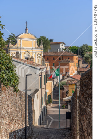 Historic street view of old Cavriana with San Biagio church Italian flag pastel facades and blue sky Historic street view of old Cavriana with San Biagio church Italian flag pastel facades and blue sky 138407748