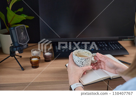 Woman holding mug at desk with monitor, keyboard, notebook and microphone Woman holding mug at desk with monitor, keyboard, notebook and microphone 138407767