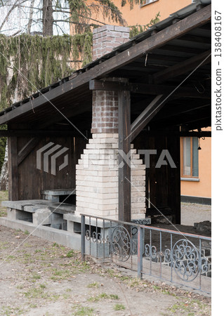 Rustic Outdoor Brick Chimney Structure With Dark Wooden Roof And Concrete Steps In Garden Setting Featuring Ornate Metal Fence Details And Mossy Trees Background 138408167
