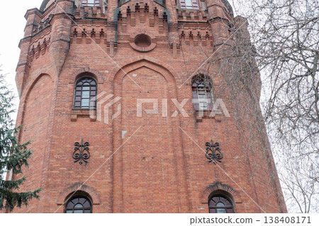 Historic Red Brick Water Tower With Gothic Arched Windows And Ornate Metal Decorations Set Against Bare Winter Trees Under Overcast Sky Historic Red Brick Water Tower With Gothic Arched Windows And Ornate Metal Decorations Set Against Bare Winter Trees Under Overcast Sky 138408171