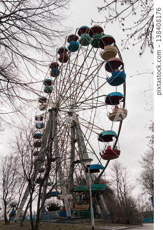 Vintage Colorful Ferris Wheel Amidst Bare Trees In Overcast Amusement Park Scene With Red Blue And Green Passenger Cars Standing Tall Against Gray Sky Vintage Colorful Ferris Wheel Amidst Bare Trees In Overcast Amusement Park Scene With Red Blue And Green Passenger Cars Standing Tall Against Gray Sky 138408176